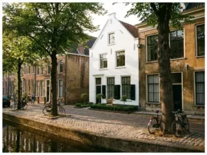 Wit herenhuis met Te Koop bord tussen oudere huizen aan grachtenstraat, gouden middaglicht op kasseien